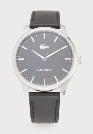 Montre Lacoste avec un boîtier en acier inoxydable argenté, un cadran gris, une aiguille des secondes bleue et un bracelet en cuir noir avec des détails de couture.