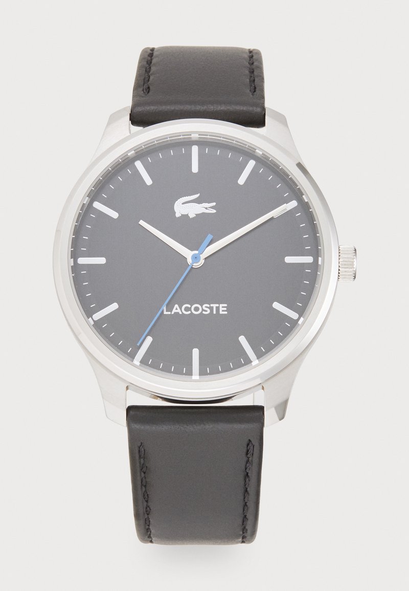 Lacoste laikrodis su sidabriniu nerūdijančio plieno korpusu, pilku ciferblatu, mėlyna antrine rodykle ir juodu odiniu laikrodžio dirželiu su siuvinėjimu.