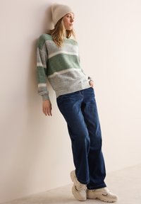 Grå och grön randig tröja, lösa blå jeans, vita sneakers och en beige rippad beanie, lutad mot en vägg.