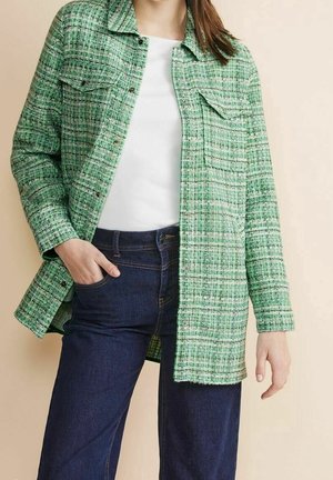 Frau trägt grüne karierte Jacke mit Knöpfen über weißem Hemd und dunkelblauen Jeans, steht mit einer Hand in der Tasche vor beigem Hintergrund.