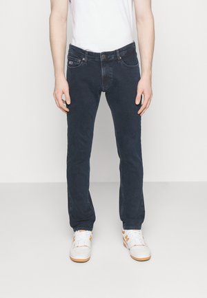 Hombre vestido con vaqueros ajustados azul oscuro, camiseta blanca y zapatillas blancas con detalles en naranja, de pie contra un fondo claro y liso.