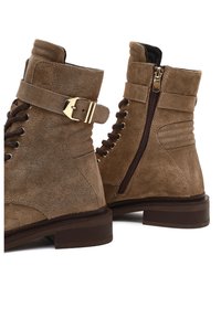 Bottes chevilles en suede marron avec une fermeture éclair sur le côté, un laçage sur le devant, un accent en boucle dorée et une semelle légèrement effilée marron.