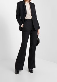 Černý oblek s přiléhavým sakem a rozšířenými kalhotami. Tkanina se jeví jako hladká, s mírným leskem. Nosený s světlým topem a kotníkovými botami.