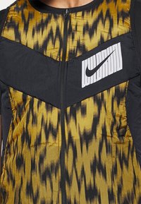 Weste mit Steppung in einem Mix aus goldenem Gelb und schwarzen geometrischen Mustern, mit einem schwarzen Nike-Logo auf der Brust und einem Reißverschluss.