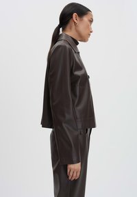 Braune Kunstleder-Cropped-Jacke mit kragenlosem Ausschnitt, Seitentaschen und glatter Textur, getragen über passenden Hosen.