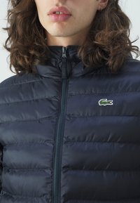 Marinblå lätt pufferjacka med horisontella sömmar, hög krage och framdragkedja. Har en liten broderad grön krokodil-logotyp.