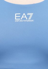 Kék sportos ujjatlan felső sima anyagból, kerek nyakkivágással és a mellrészen fehér "EA7 EMPORIO ARMANI" nyomott logóval.