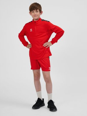 Ragazzo in piedi con le mani sui fianchi, indossa abbigliamento sportivo rosso, calzini bianchi e scarpe da ginnastica nere su uno sfondo chiaro e semplice.