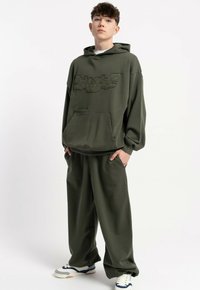 Oliwkowa bluza z kapturem z teksturowanym logo, kieszenią z przodu i sznurkiem do ściągania; w zestawieniu z luźnymi oliwkowymi spodniami i białymi butami sportowymi.