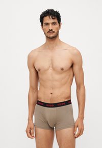 Culotte beige avec une coupe ajustée et une ceinture noire arborant le logo « HUGO » en rouge. Tissu doux et sans coutures extérieures.