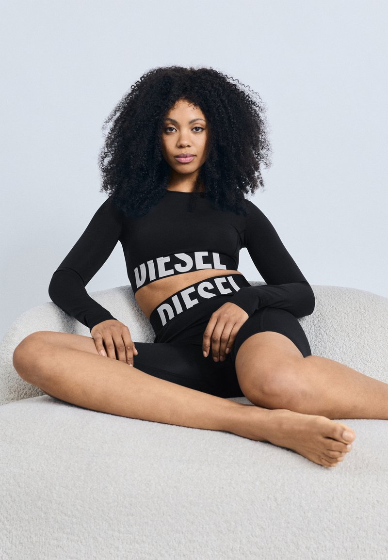 Czarny długi rękaw crop top i dopasowane szorty z napisem "DIESEL" w grubych białych literach. Modelka siedzi na jasnej, fakturowanej sofie.