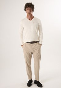 Chemise crème à manches longues en V avec un petit logo, associée à un pantalon beige clair et des bottines noires.