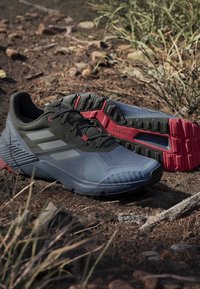 Buty do biegania w terenie w ciemnoszarym i niebieskim kolorze, z teksturowaną cholewką, trzema białymi paskami oraz czerwonym akcentem na podeszwy. Design skoncentrowany na przyczepności.