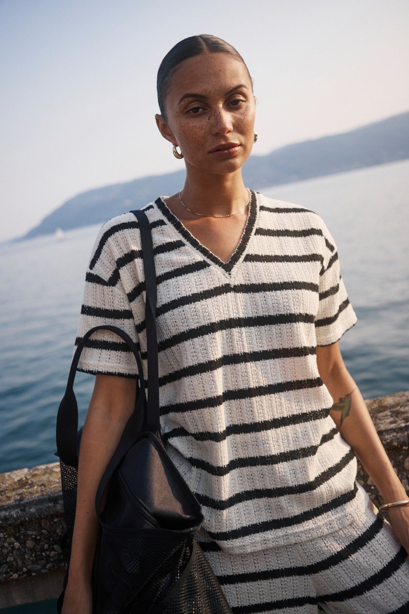 Ribaštruktuuriga kudum valges ja mustas, millel on V-kaelus ja lühikesed varrukad. Kandev must kott, mereäärse taustaga.