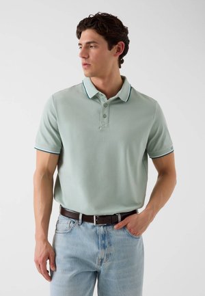 Homme portant un polo vert clair à manches courtes avec une bordure foncée, rentré dans un jean bleu clair avec une ceinture marron foncé, debout devant un fond uni.