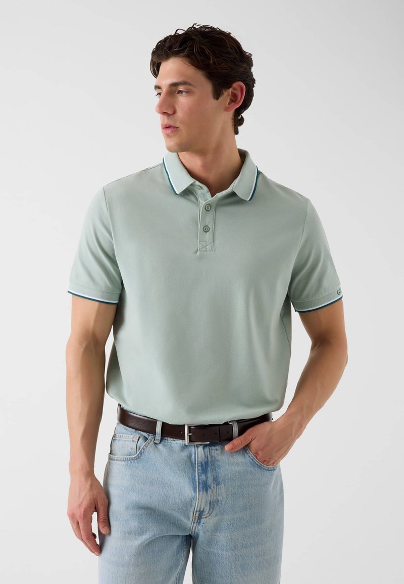 Homme portant un polo vert clair à manches courtes avec une bordure foncée, rentré dans un jean bleu clair avec une ceinture marron foncé, debout devant un fond uni.