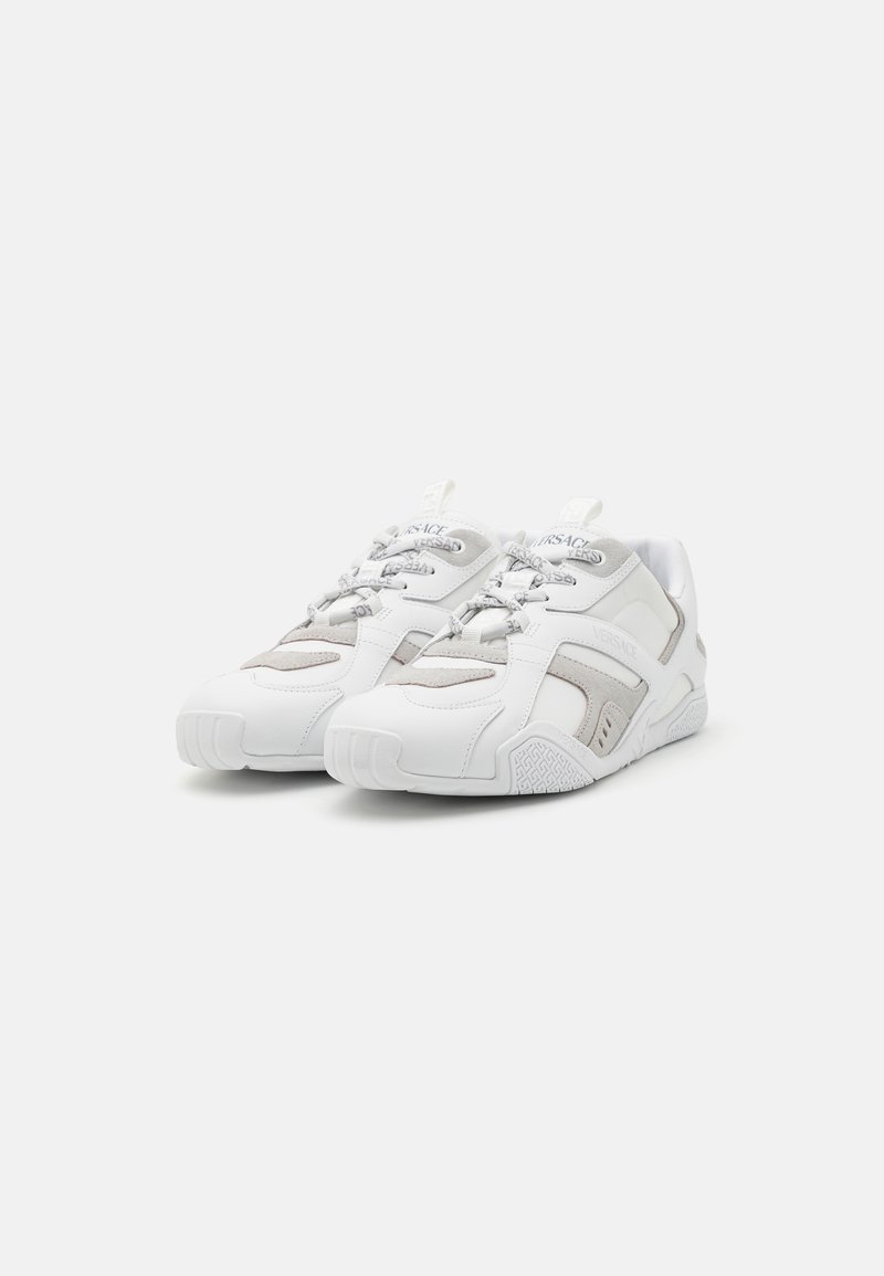 White sneakers featuring a mix of smooth and textured materials, with grey accents, a rounded toe, and distinctive lace design.