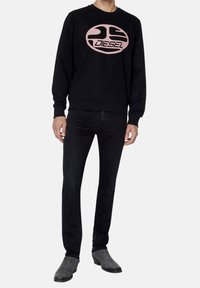 Sudadera negra con un logo rosa que dice "DIESEL," combinada con jeans negros y unos zapatos grises texturizados, mostrando un atuendo casual.