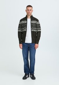 Mørk grønn strikket cardigan med glidelås, med hvite geometriske mønstre på armene. Kombinert med blå jeans og svarte joggesko.
