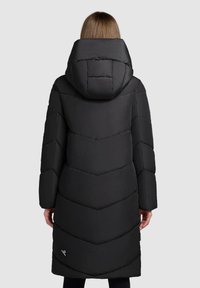 Person, der einen langen schwarzen Daunenmantel mit großer Kapuze trägt, steht mit dem Rücken zu einer schlichten Hintergrund.