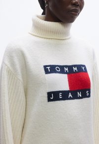 Elefántcsont színű pulóver magas bordázott nyakkal, elöl piros és kék "TOMMY JEANS" logóval. Puha, texturált anyagból, széles ujjakkal.