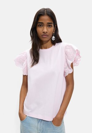 Kobieta o długich ciemnych włosach nosi jasnoróżową bluzkę z aksamitnymi koronkowymi bufiastymi rękawami i jasnoniebieskie jeansy, stoi z rękami w kieszeniach.