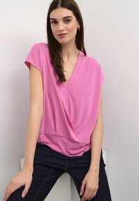 Rosa, kortärmad topp med V-ringning, som har en inslagning design och slät textur. I kombination med mörka jeans, sitter mot en neutral bakgrund.