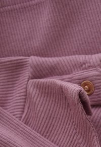 Close-up van stof in stofroze met ribbels, met een omgevouwen manchet en een enkele koperen drukknoop.