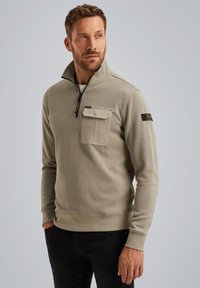 Suéter beige claro con cuello con cremallera, textura acanalada y un bolsillo en el pecho. Presenta un parche con el logo en la manga izquierda. Combinado con pantalones oscuros.