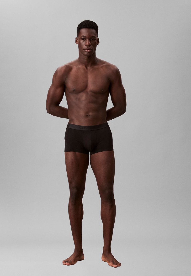 Boxer briefs negros en una forma ajustada, hechos de una tela suave con una cinturilla elástica. El modelo está de pie con los brazos detrás de la espalda, sobre un fondo neutro.