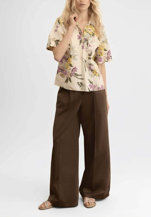 Donna con blusa beige a maniche corte floreale, pantaloni larghi marrone scuro e sandali beige, in piedi davanti a uno sfondo chiaro e semplice.