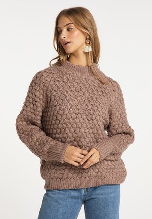 Frau schaut zur Seite, trägt einen strukturierten braunen Strickpullover, blaue Jeans und Quastenohrringe, die Hände locker in der Taille verschränkt.