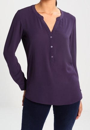 Kobieta ubrana w długorękawową, ciemnofioletową bluzkę z dekoltem w serek i trzema guzikami, zestawioną z ciemnymi spodniami na jednolitym tle.