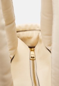 Beżowa skórzana torba na ramię ze złotym suwakiem, ukazująca miękką teksturę, gładkie wykończenie oraz widoczny suwak z logo przy otwarciu.