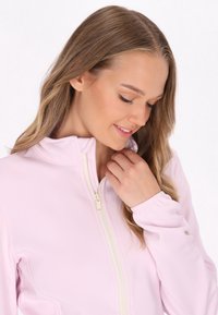 Világos rózsaszín kabát magas gallérral, cipzáras záródással és rugalmas mandzsettákkal; sima textúrájú, minimális varrással és minták nélkül.