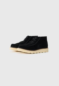 Black suede ankle boots with beige crepe soles and black laces, side-by-side on a plain light gray background.