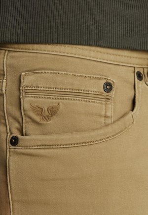Beige kavbojke iz denima vsebujejo majhno, vezeno krilato logo na sprednjem žepu, s kontrastnim temnim šivanjem in črnimi gumbi.