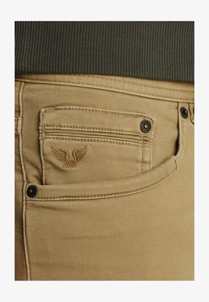 Beige denim jeans har et lille, broderet vinge-logo på frontlommen, med kontrasterende mørk syning og sorte knapdetaljer.