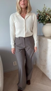 Woman wearing a cream buttoned cardigan and gray pinstriped wide-leg trousers, standing indoors next to a vase with white and red flowers.