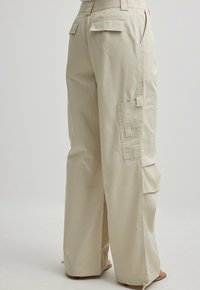 Beige cargo-housut, joissa on löysä istuvuus, useita sivutaskuja, napin yksityiskohtia ja sileä, puuvillamainen rakenne. Mukana myös takataskujen läpät.