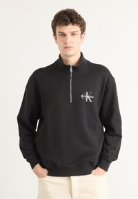 Musta vetoketjuhuppari, joka on valmistettu pehmeästä kankaasta. Sen ominaisuuksiin kuuluu korkea kaulus, ribbaatut hihansuut ja hopeinen Calvin Klein -logo rinnassa.