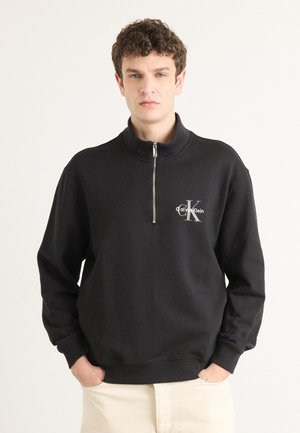 Hanorac negru cu fermoar, fabricat dintr-un material moale, având un guler înalt, manșete ribbate și un logo argintiu Calvin Klein pe piept.