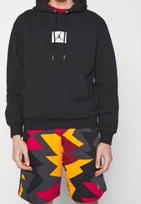 Persona che indossa una felpa nera con cappuccio con il logo Jumpman bianco e pantaloncini dai colori vivaci con motivi geometrici frastagliati in rosso, giallo, grigio e nero.