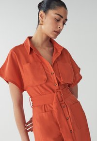 Rust-colored shirt dress with short sleeves, two chest pockets, button front, and a fabric belt. Smooth texture and relaxed fit.