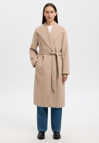 Kvinne står iført en beige ullfrakk med belte, blå jeans, svarte sko, og en hvit skjorte mot en ensfarget hvit bakgrunn.
