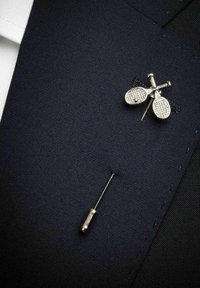 Silverfärgad reversnål i form av korslagda tennisracketar, fästad på reversen av den mörkblå kostymjackan ovanför en silverfärgad nål.