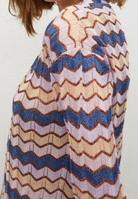 Pull en tricot avec motif en zigzag dans des couleurs pastel : beige, bleu et lavande. Présente des accents métalliques et une finition texturée.