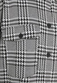 Tissu à motif pied-de-poule noir et blanc avec une poche à rabat fermée par un bouton et deux boutons noirs visibles supplémentaires.