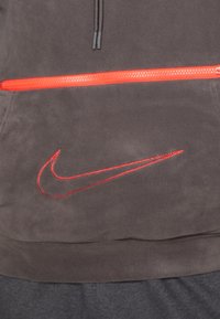 Felpa con cappuccio grigio scuro in pile, dotata di una tasca frontale con zip, caratterizzata da una zip rossa brillante. Il logo Nike è ricamato in rosso sulla parte anteriore.