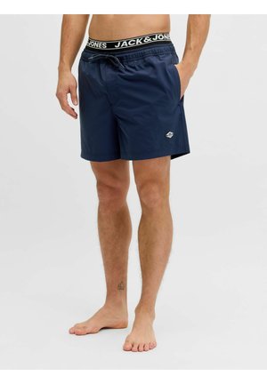 Mężczyzna noszący granatowe kąpielówki z bocznymi kieszeniami i paskiem z logo "Jack & Jones", stojący boso na jednolitym tle.